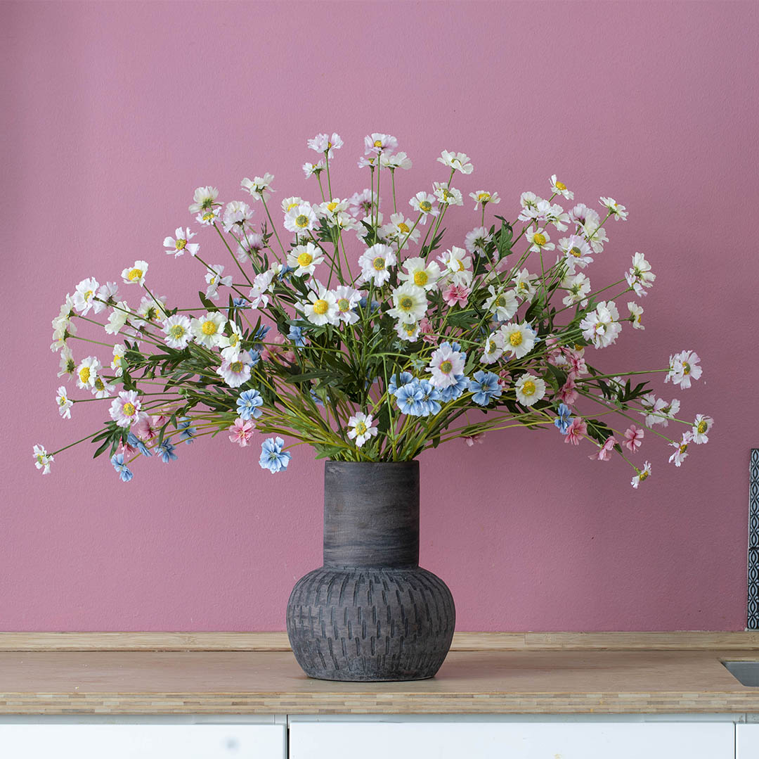 En bred bukett med vita och rosa blommor i en mörkgrå kruka framför en rosa vägg.